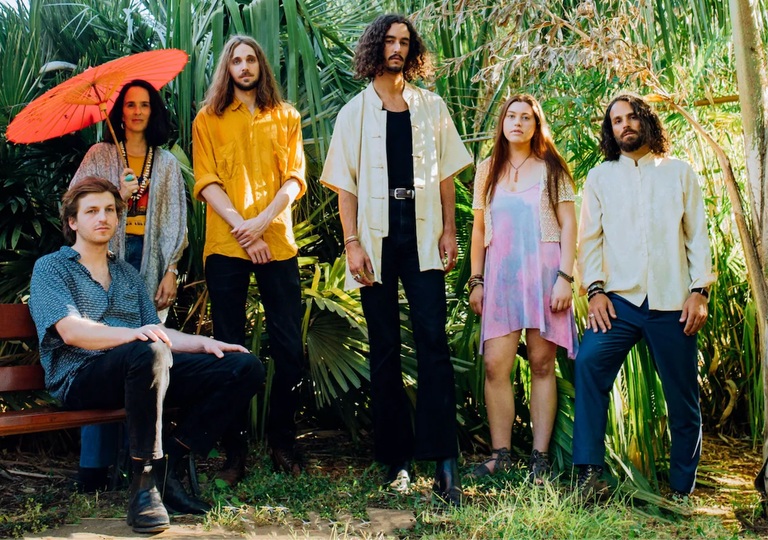 Adelaide psychedelic rock collective Sons of Zöku posing outdoors surrounded by lush green tropical foliage and holding a red parasol.
